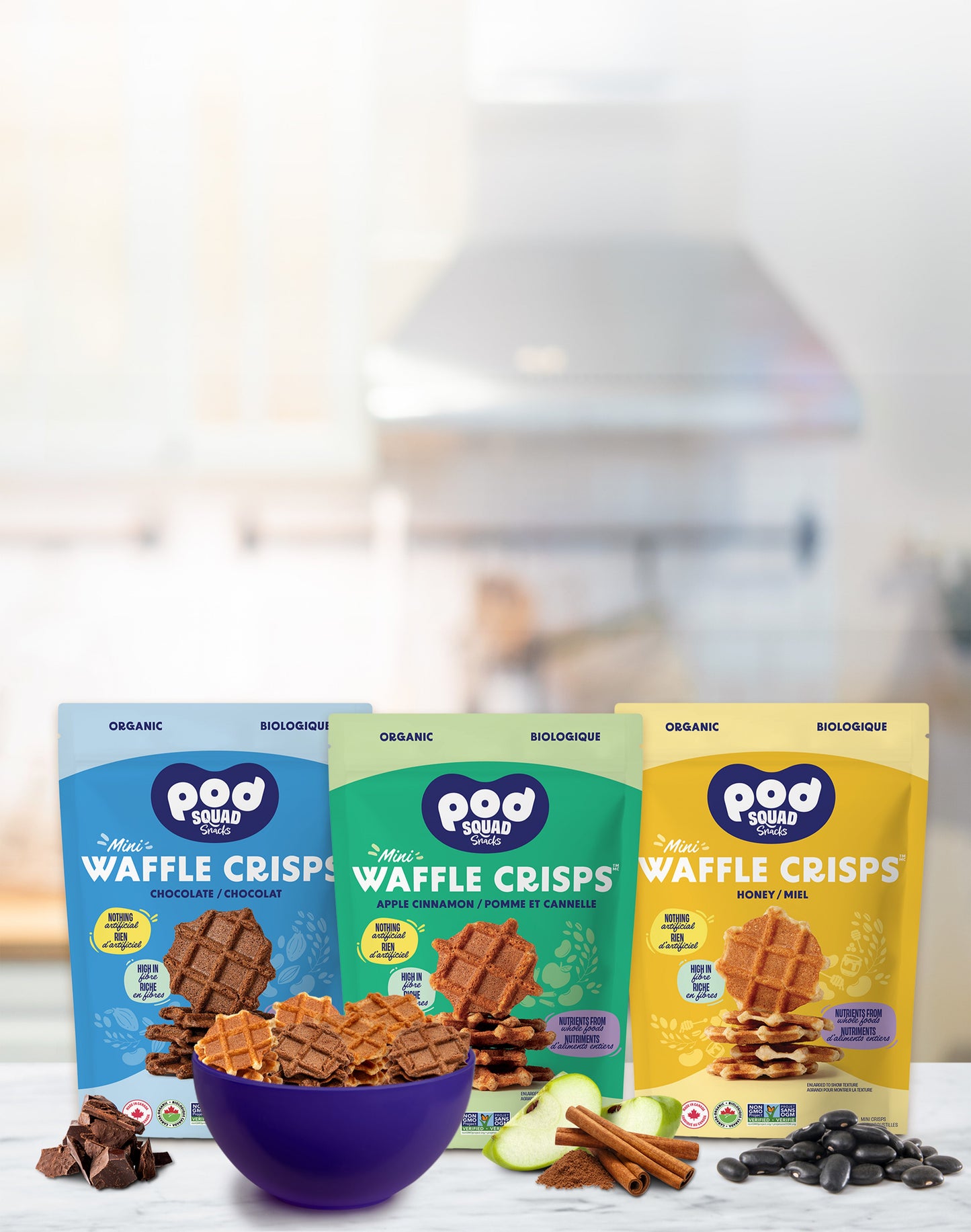 Three bags of Pod Squad Snacks Mini Waffle Crisps with a bowl of waffle crisps and ingredients on a blurred background.